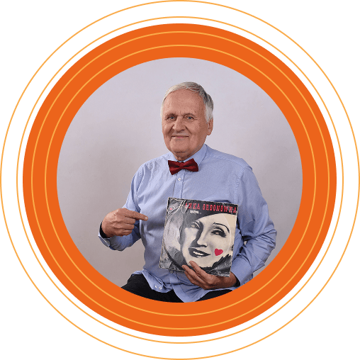 Starszy mężczyzna w błękitnej koszuli i bordowej muszce trzyma winylową płytę z wizerunkiem Hanki Ordonówny. Uśmiecha się i wskazuje na okładkę.